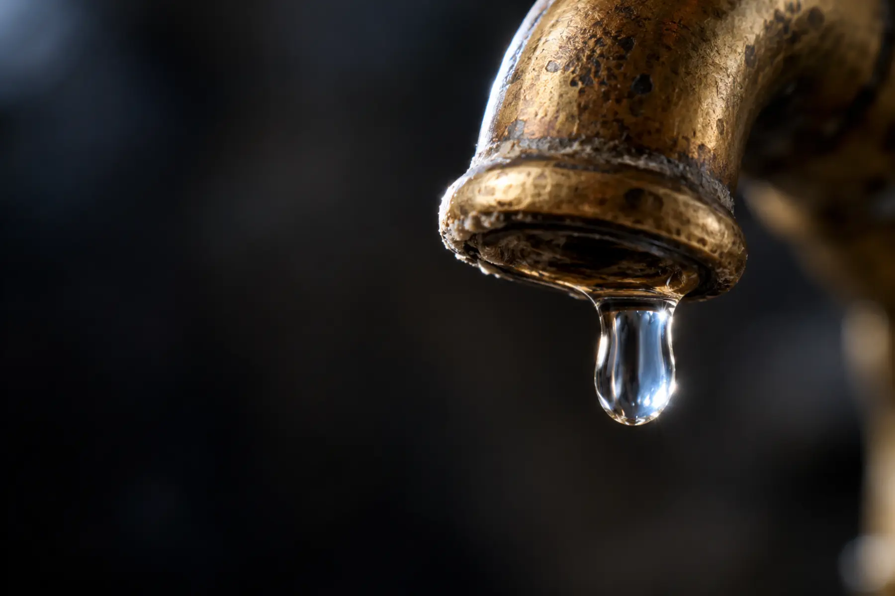 A single drop of water suspended from the spout of a weathered brass tap, backlit against a dark background